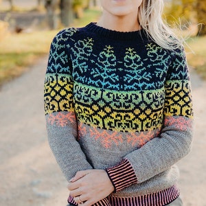 May include: A woman wearing a colorful knitted sweater with a geometric pattern. The sweater has a dark blue base with a rainbow gradient and a gray bottom. The woman is standing in a path with trees in the background.