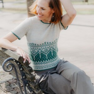 May include: A short-sleeved knit top featuring a cream-colored upper section and a green lower section with a detailed geometric pattern. The garment has a round neckline and green trim along the sleeve edges and the bottom hem of the shirt.