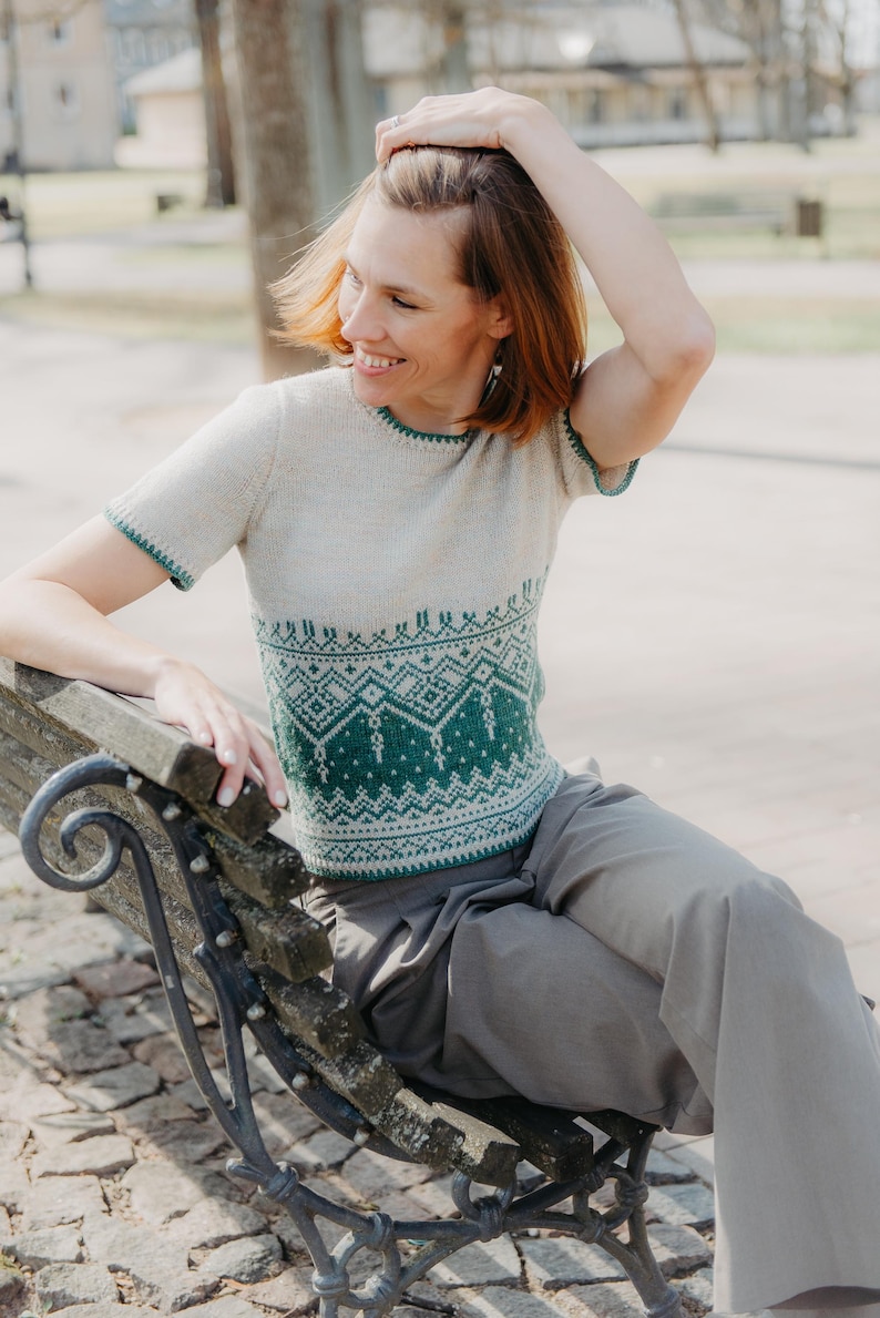 May include: A short-sleeved knit top featuring a cream-colored upper section and a green lower section with a detailed geometric pattern. The garment has a round neckline and green trim along the sleeve edges and the bottom hem of the shirt.