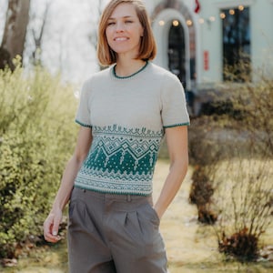 May include: A short-sleeved knit sweater featuring a cream-colored upper section and a dark teal lower section with a white geometric pattern. The garment has a round neckline and matching dark teal trim on the collar and sleeve cuffs.