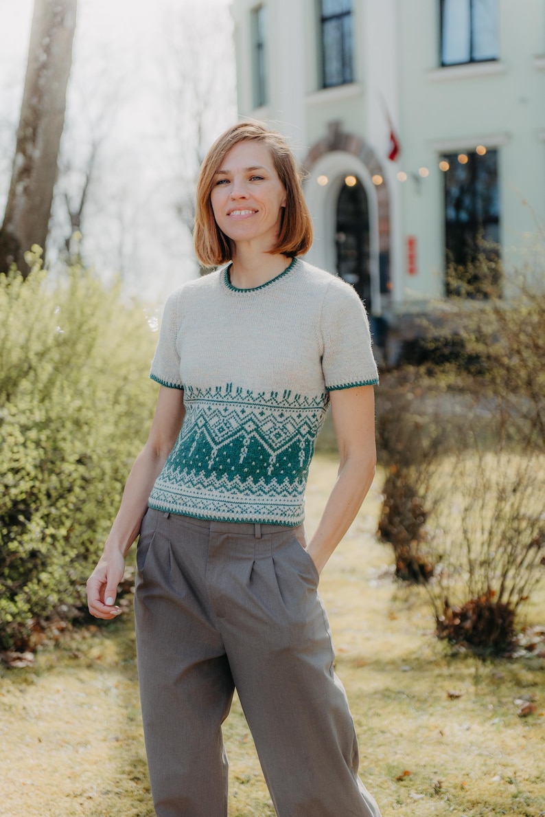 May include: A short-sleeved knit sweater featuring a cream-colored upper section and a dark teal lower section with a white geometric pattern. The garment has a round neckline and matching dark teal trim on the collar and sleeve cuffs.