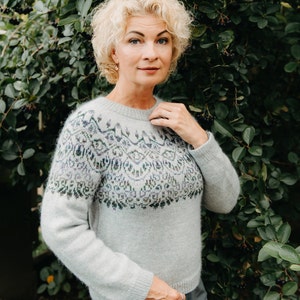 May include: A light blue sweater with a patterned yoke featuring geometric designs in shades of blue, green, and purple. The sweater has long sleeves and a crew neck. The background is a lush green bush.