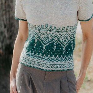 May include: A short-sleeved knit sweater featuring a cream-colored upper section and a dark green lower section with an intricate geometric pattern. The garment has a round neckline and sleeve cuffs trimmed in dark green, paired with grey trousers.