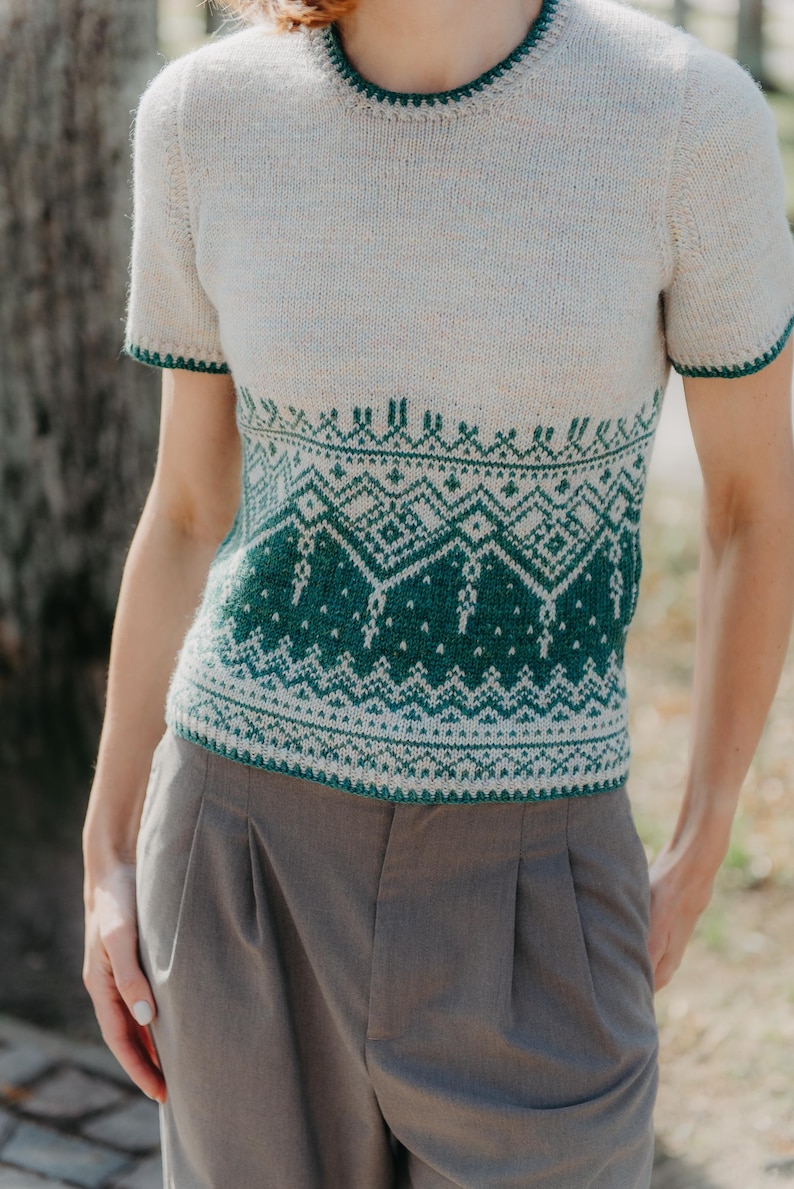 May include: A short-sleeved knit sweater featuring a cream-colored upper section and a dark green lower section with an intricate geometric pattern. The garment has a round neckline and sleeve cuffs trimmed in dark green, paired with grey trousers.