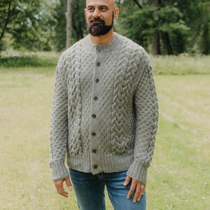 Peut inclure: Un cardigan gris en tricot torsadé avec une fermeture boutonnée. Le pull a un motif texturé et est porté avec un jean bleu. La personne se tient dans un champ herbeux avec des arbres en arrière-plan.