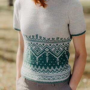 May include: A short-sleeved knit sweater featuring a cream-colored upper section and a dark green lower section with a detailed geometric pattern. The garment has a round neckline and matching dark green trim on the sleeve cuffs and collar.