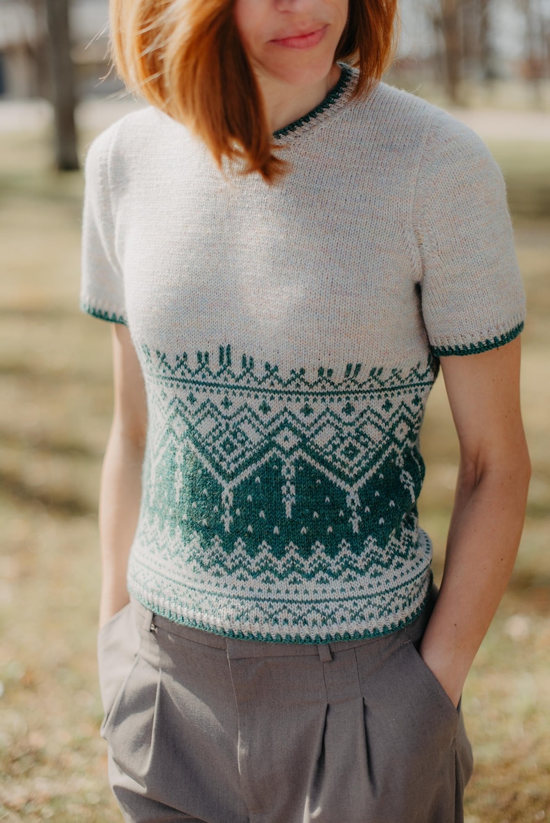 May include: A short-sleeved knit sweater featuring a cream-colored upper section and a dark green lower section with a detailed geometric pattern. The garment has a round neckline and matching dark green trim on the sleeve cuffs and collar.