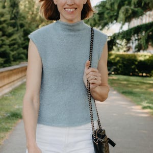 Puede incluir: Un suéter sin mangas de color azul claro con cuello alto y textura difusa, combinado con pantalones blancos y un pequeño bolso bandolera negro acolchado con cadena dorada. El suéter tiene un tejido sencillo y dobladillo recto.
