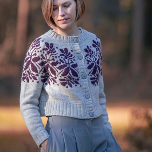 Può includere: Una donna indossa un cardigan grigio con un motivo floreale viola e pantaloni grigi. Il cardigan ha una chiusura con bottoni.