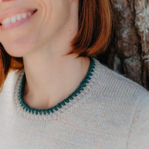 May include: A light beige knit sweater featuring a round neckline with a contrasting dark green crocheted trim. The sweater has a simple, clean design with a smooth texture and a ribbed collar edge that highlights the dark green yarn detail.