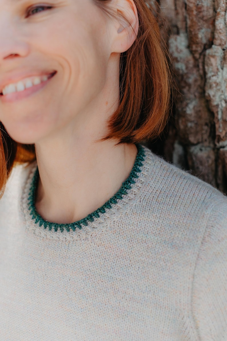 May include: A light beige knit sweater featuring a round neckline with a contrasting dark green crocheted trim. The sweater has a simple, clean design with a smooth texture and a ribbed collar edge that highlights the dark green yarn detail.