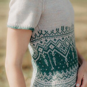 May include: A short-sleeved knit sweater featuring a cream-colored base with a detailed green geometric pattern across the midsection and hem. The garment has a round neckline and ribbed trim on the sleeves and bottom edge in a matching green.