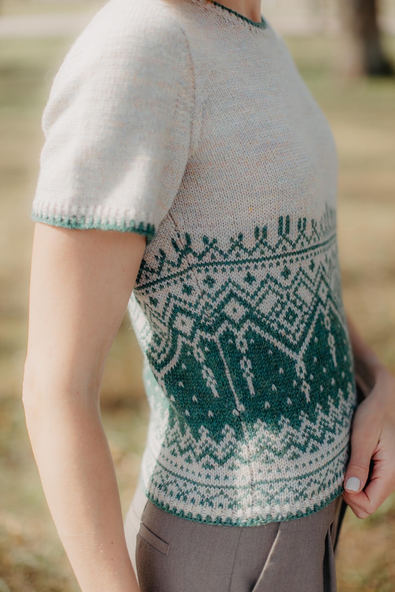 May include: A short-sleeved knit sweater featuring a cream-colored base with a detailed green geometric pattern across the midsection and hem. The garment has a round neckline and ribbed trim on the sleeves and bottom edge in a matching green.