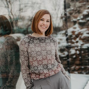 May include: A woman wearing a patterned sweater with a diamond design in shades of brown, gray, and burgundy. She is also wearing gray pants. The background includes a reflective surface and a snowy outdoor scene.