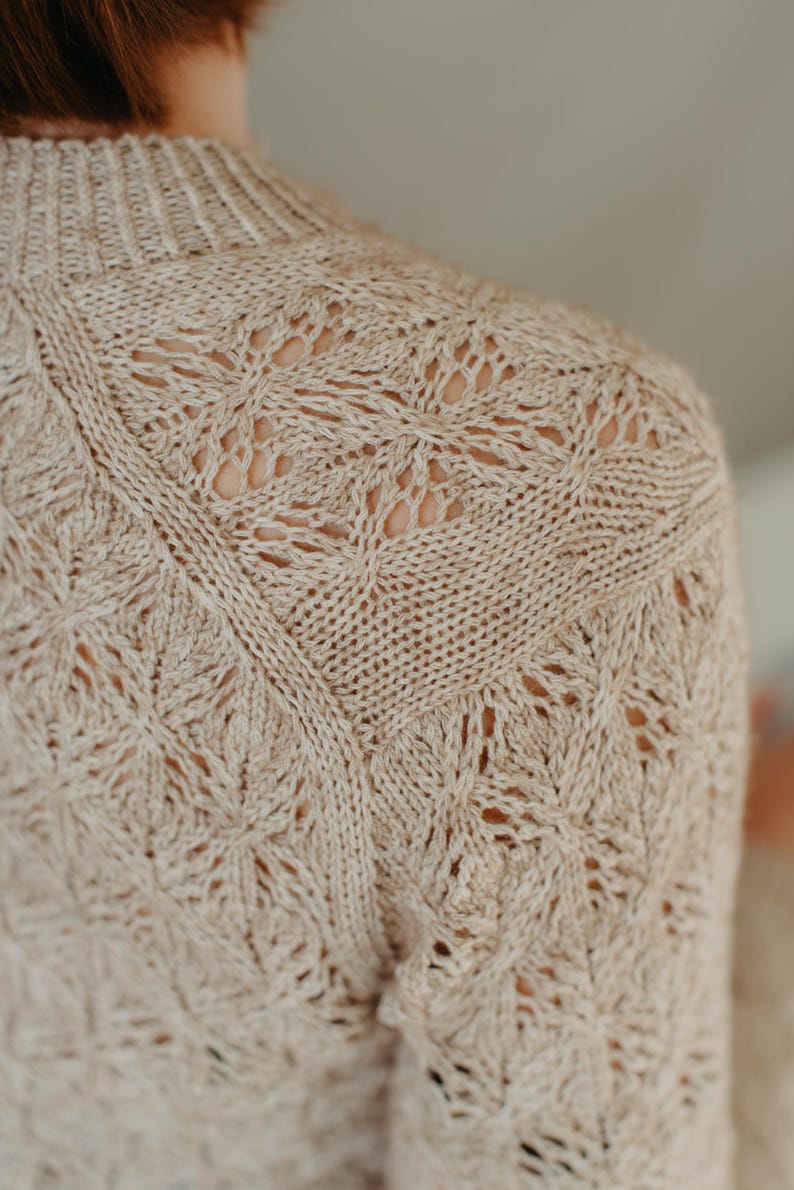 May include: Close-up of a beige knitted sweater with intricate openwork detailing. The sweater features a crew neck and a textured pattern across the shoulders and upper back. The knitwear is a neutral color, suitable for various outfits.