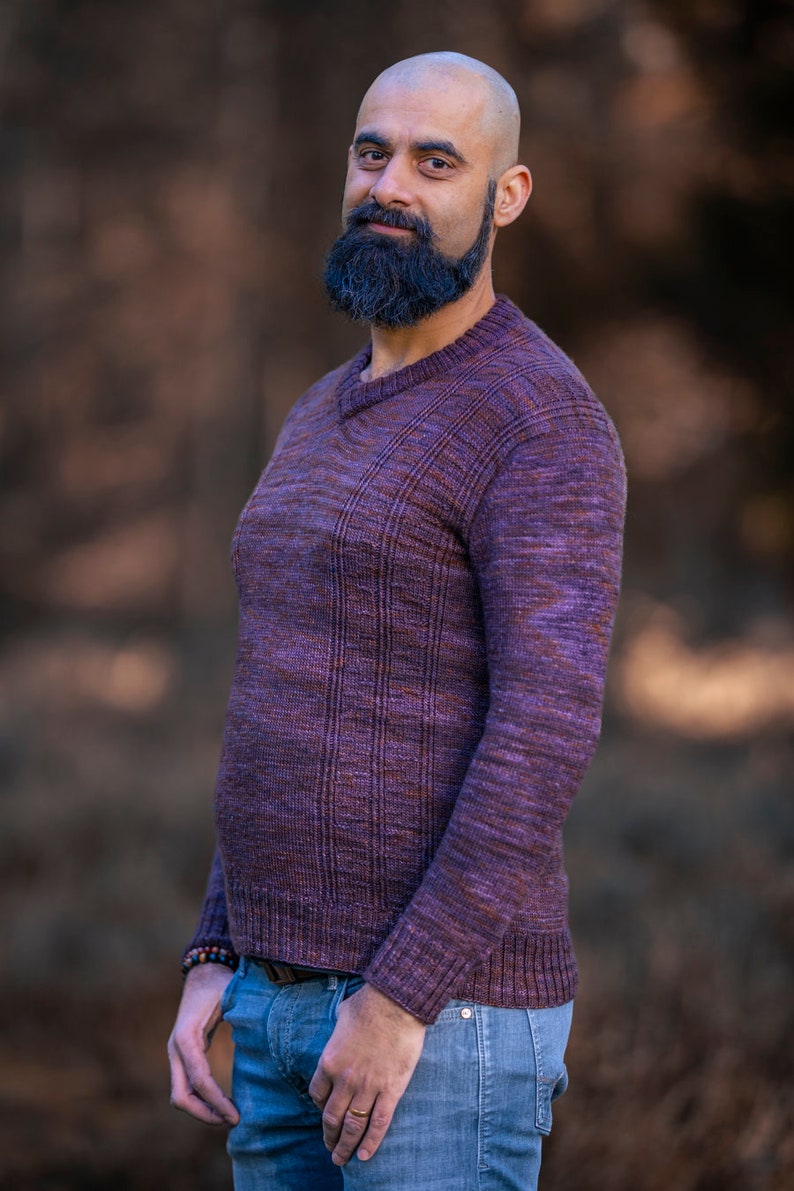 May include: A man with a shaved head and a dark beard is wearing a purple sweater with a v-neck and blue jeans.