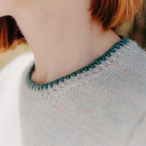 May include: A light-colored, speckled knit sweater featuring a round neckline with a contrasting dark green crocheted trim. The sweater is crafted from a textured yarn with subtle flecks of color throughout the fabric, creating a soft appearance.