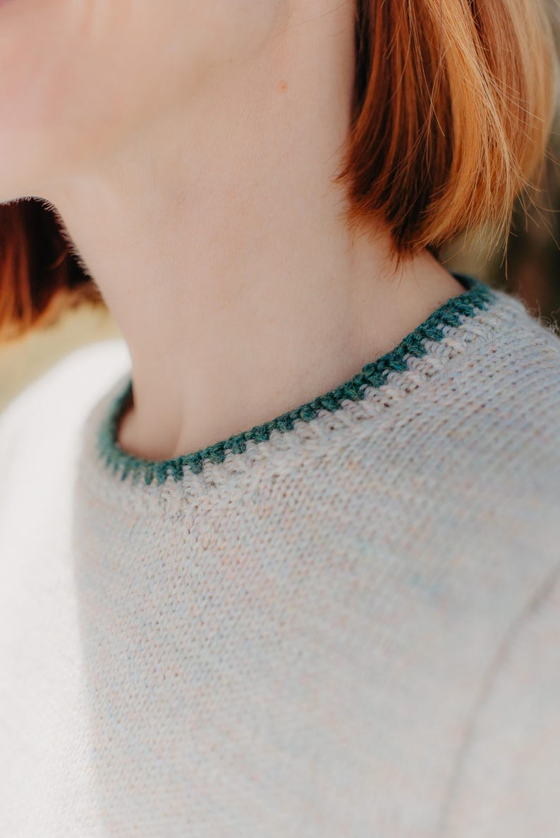 May include: A light-colored, speckled knit sweater featuring a round neckline with a contrasting dark green crocheted trim. The sweater is crafted from a textured yarn with subtle flecks of color throughout the fabric, creating a soft appearance.
