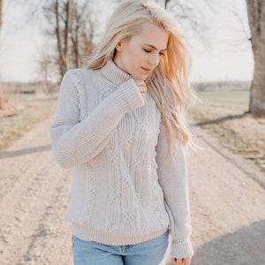 May include: A woman wearing a light beige cable knit sweater with a turtleneck and blue jeans. She is standing on a dirt road with trees in the background.