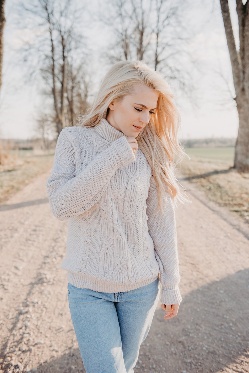 May include: A woman wearing a light beige cable knit sweater with a turtleneck and blue jeans. She is standing on a dirt road with trees in the background.