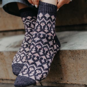 May include: A pair of patterned socks in shades of navy blue and pale pink. The socks feature a repeating geometric design. The cuffs, heels, and toes are a solid navy blue. The socks are made of a soft, knitted material.