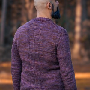May include: A man wearing a purple and brown sweater with a ribbed texture. He is standing in a forest with trees in the background.