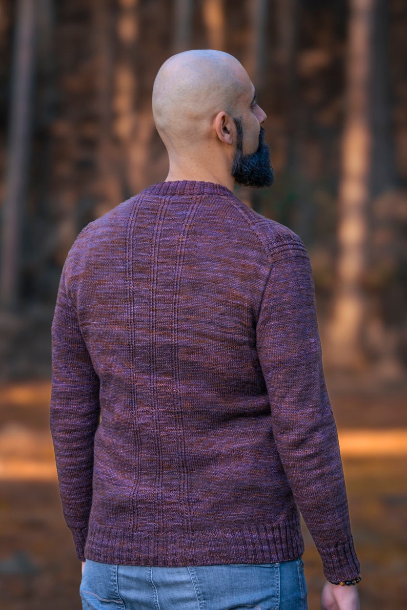 May include: A man wearing a purple and brown sweater with a ribbed texture. He is standing in a forest with trees in the background.