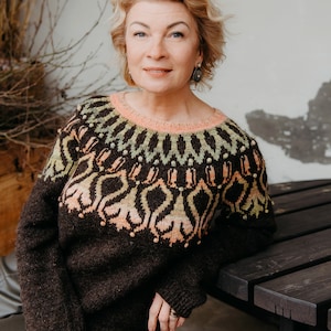 May include: A woman wearing a dark brown sweater with a patterned design in shades of peach, green, and cream. The sweater has a round neckline and long sleeves. The woman is sitting on a dark wooden bench.