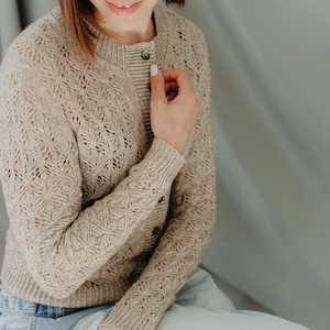 Puede incluir: Cárdigan beige de punto calado con cierre de botones. El suéter presenta un delicado patrón calado y puños acanalados. La modelo lleva vaqueros azul claro. El cárdigan está hecho de un material suave y texturizado.