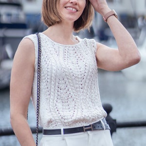 Op de afbeelding: Een vrouw draagt een wit, gehaakt, mouwloos topje met een zwarte riem en een witte broek. Ze houdt haar hand tegen haar hoofd en glimlacht.