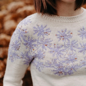 以下が含まれることがあります： 紫色の花柄の白いニットセーター。セーターはクルーネックで長袖です。