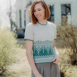 May include: A short-sleeved knit sweater featuring a cream-colored upper section and a dark green lower section adorned with a detailed geometric pattern. The garment has a round neckline and green trim along the collar and sleeve edges.