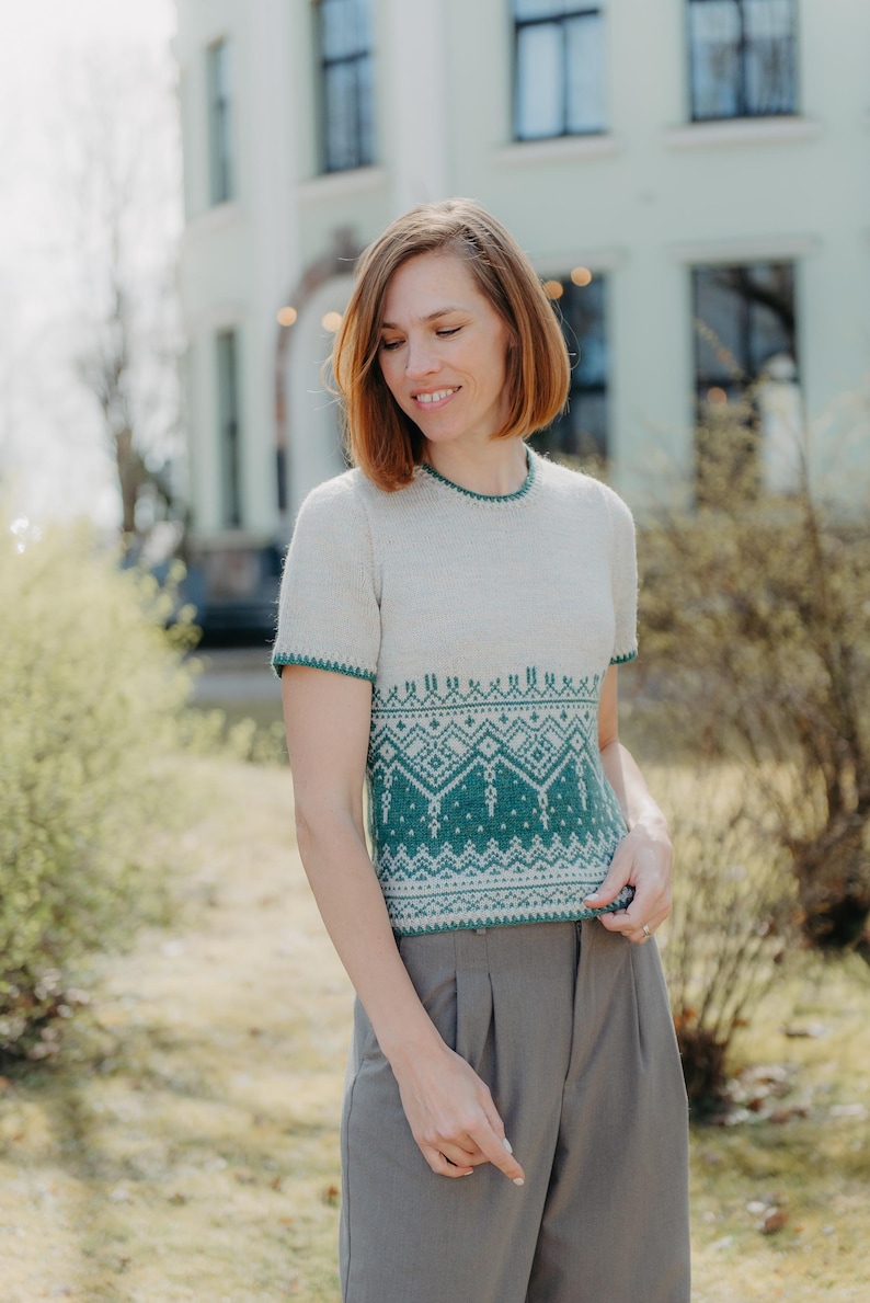 May include: A short-sleeved knit sweater featuring a cream-colored upper section and a dark green lower section adorned with a detailed geometric pattern. The garment has a round neckline and green trim along the collar and sleeve edges.