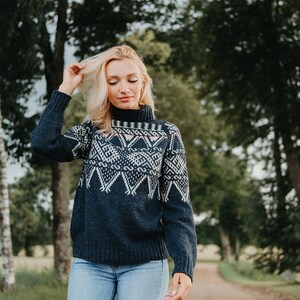 Op de afbeelding: Een vrouw draagt een marineblauwe trui met een wit geometrisch patroon en een coltrui. De trui wordt gecombineerd met een lichtblauwe jeans. De achtergrond toont een zandpad en bomen.
