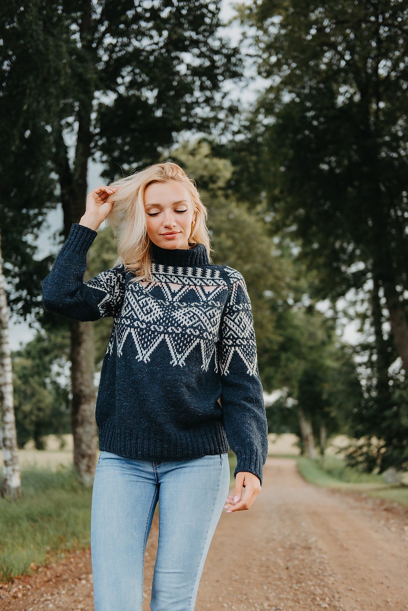 Op de afbeelding: Een vrouw draagt een marineblauwe trui met een wit geometrisch patroon en een coltrui. De trui wordt gecombineerd met een lichtblauwe jeans. De achtergrond toont een zandpad en bomen.
