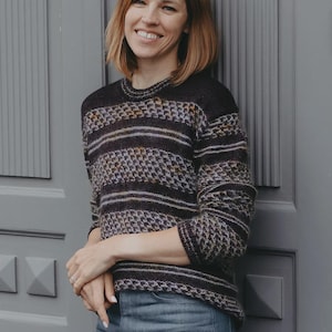 Pode incluir: Um suéter escuro com listras horizontais e padrões texturizados. O suéter tem gola redonda e mangas compridas. A pessoa está usando jeans azuis. O fundo é uma porta cinza.