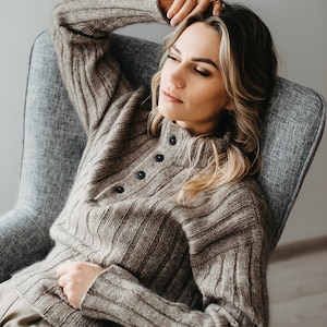 以下が含まれることがあります： 灰色のアームチェアに座っている、ボタン留めのブラウンニットセーターを着た女性