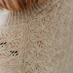 May include: Close-up of a beige knit sweater with a high neckline. The sweater features a detailed openwork pattern with floral and geometric shapes. The texture of the knitwear is visible, showcasing the craftsmanship of the garment.