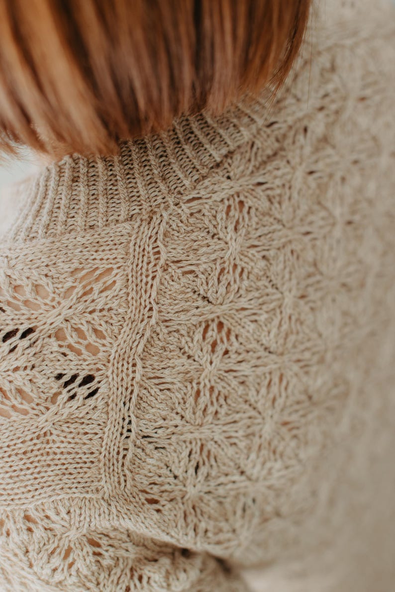 May include: Close-up of a beige knit sweater with a high neckline. The sweater features a detailed openwork pattern with floral and geometric shapes. The texture of the knitwear is visible, showcasing the craftsmanship of the garment.