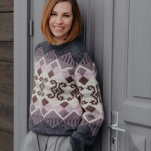 May include: A gray sweater with a geometric pattern in white, pink, and brown. The sweater has a crew neck and long sleeves. The person is leaning against a gray door.