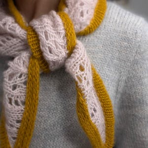 Könnte beinhalten: Ein dreieckiger Strickschal mit einem hellrosa Spitzenmuster und einem einfarbigen, senfgelben Strickrand. Der Schal ist um den Hals geknotet und zeigt die kontrastierenden Farben sowie die strukturierte Beschaffenheit des Garns.