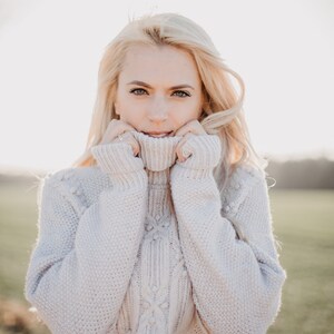 May include: A woman with blonde hair is wearing a white cable knit sweater with a turtleneck. She is holding the collar of the sweater up to her chin.