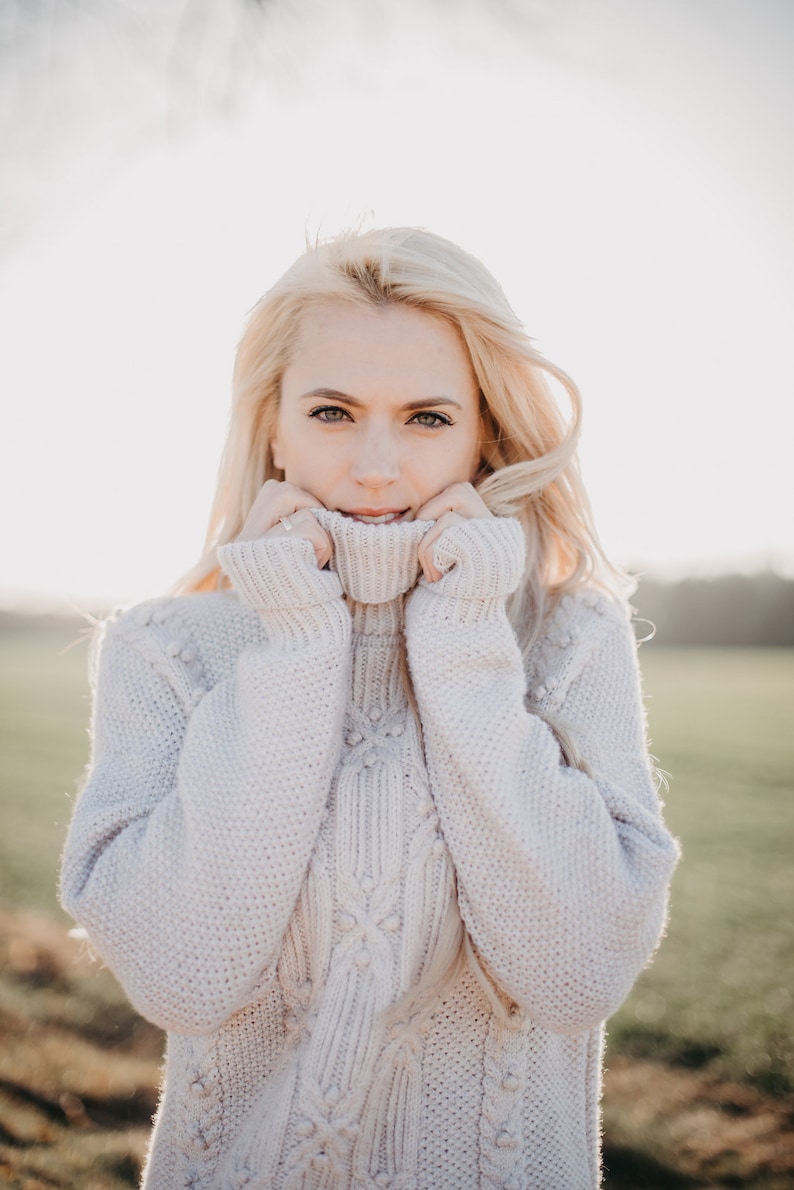 May include: A woman with blonde hair is wearing a white cable knit sweater with a turtleneck. She is holding the collar of the sweater up to her chin.