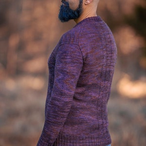 May include: A man wearing a purple and brown sweater with a ribbed texture. He is standing in a field with a blurred background.