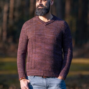 May include: A man with a shaved head and a full beard is wearing a maroon sweater with a v-neck and blue jeans. He is standing in a woodland setting with trees in the background.