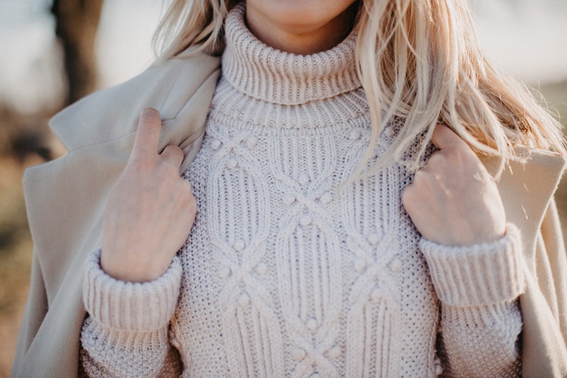May include: A close-up of a cream-colored turtleneck sweater with a cable knit pattern and small pearl-like embellishments. The sweater is worn by a person with blonde hair and is partially covered by a beige coat.