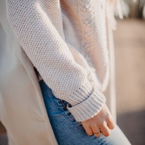 May include: A close-up of a person wearing a light beige knitted sweater with a cable knit design. The sweater is worn over a pair of blue jeans.