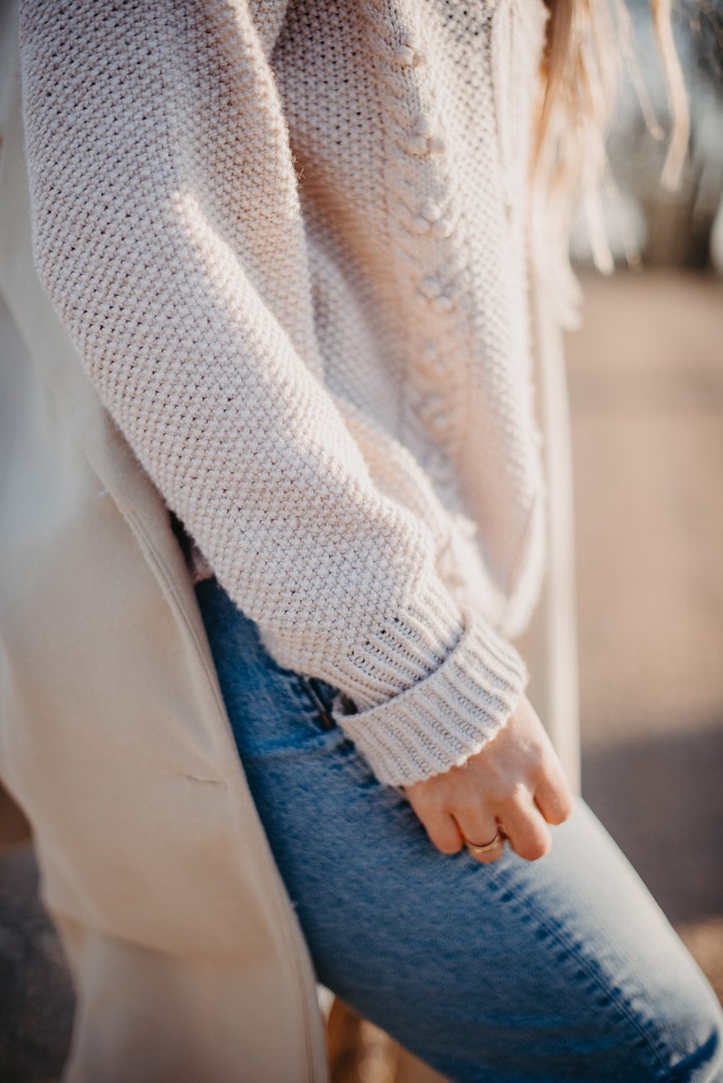 May include: A close-up of a person wearing a light beige knitted sweater with a cable knit design. The sweater is worn over a pair of blue jeans.