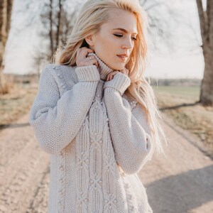May include: A woman wearing a light beige cable knit sweater with a textured pattern. The sweater has a high neck and is pulled up to her chin. She is standing in a field with trees in the background.