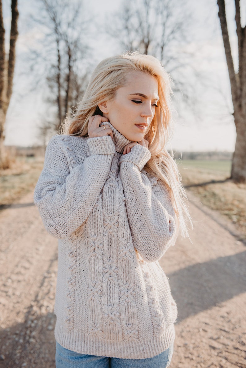 May include: A woman wearing a light beige cable knit sweater with a textured pattern. The sweater has a high neck and is pulled up to her chin. She is standing in a field with trees in the background.